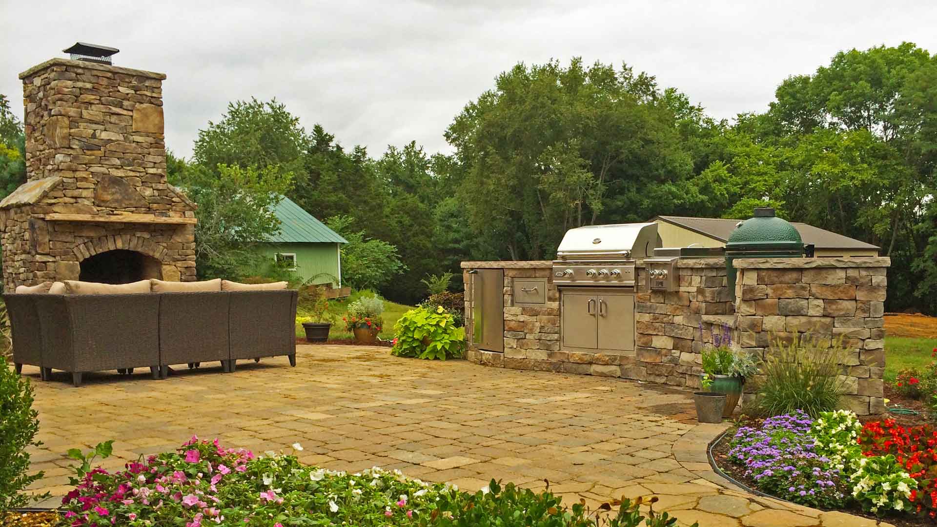 Custom stone outdoor kitchen design with a built-in grill and kamado smoker next to a matching stone fireplace on a paver patio. Patio is surrounded by gardens.