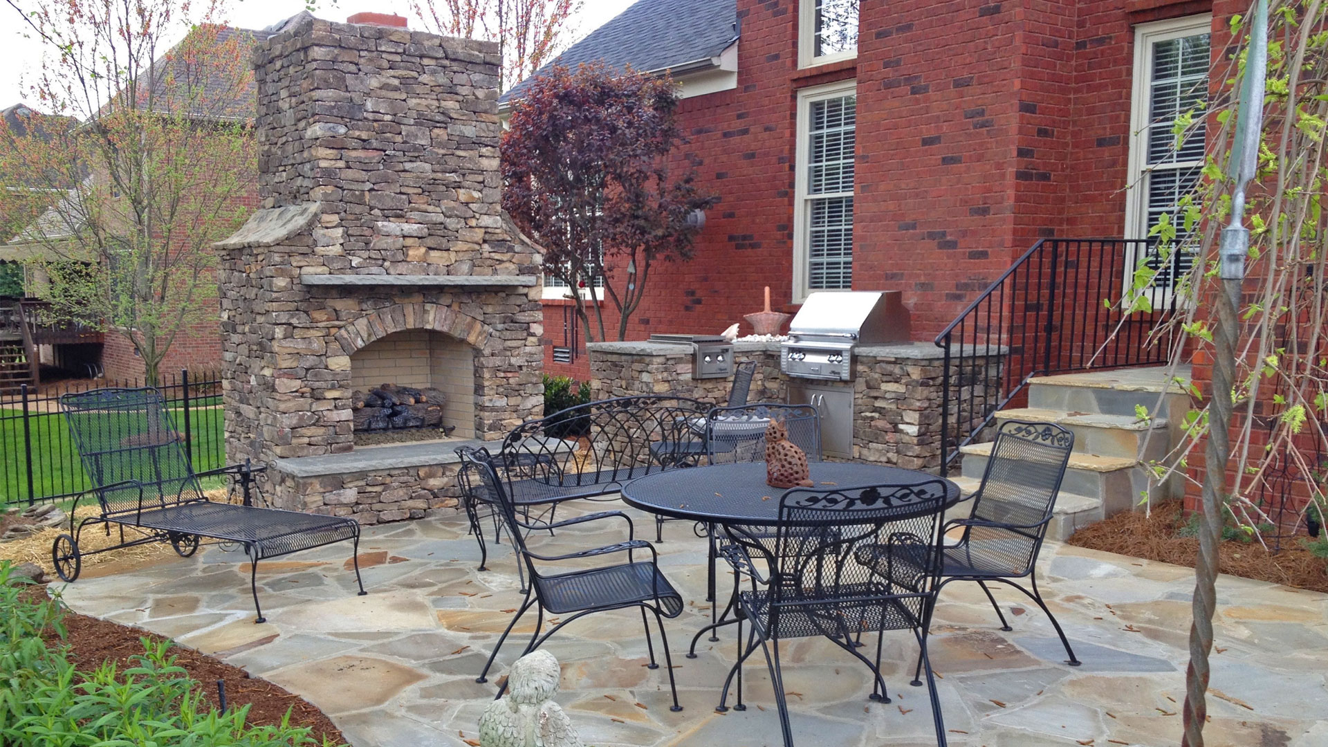 Covered outdoor kitchen and patio area with stone countertops, built in stainless steel grill, outdoor sink, and storage cabinets under a wooden pavilion, overlooking a green landscaped backyard.