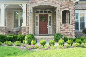 A welcoming front entrance with a red door framed by stone and brickwork, surrounded by neatly trimmed shrubs, small boxwoods, and blooming pink flowers along the walkway.