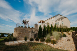A large stone retaining wall supporting an elevated patio, bordered with evergreen trees, ornamental grasses, and river rock landscaping.