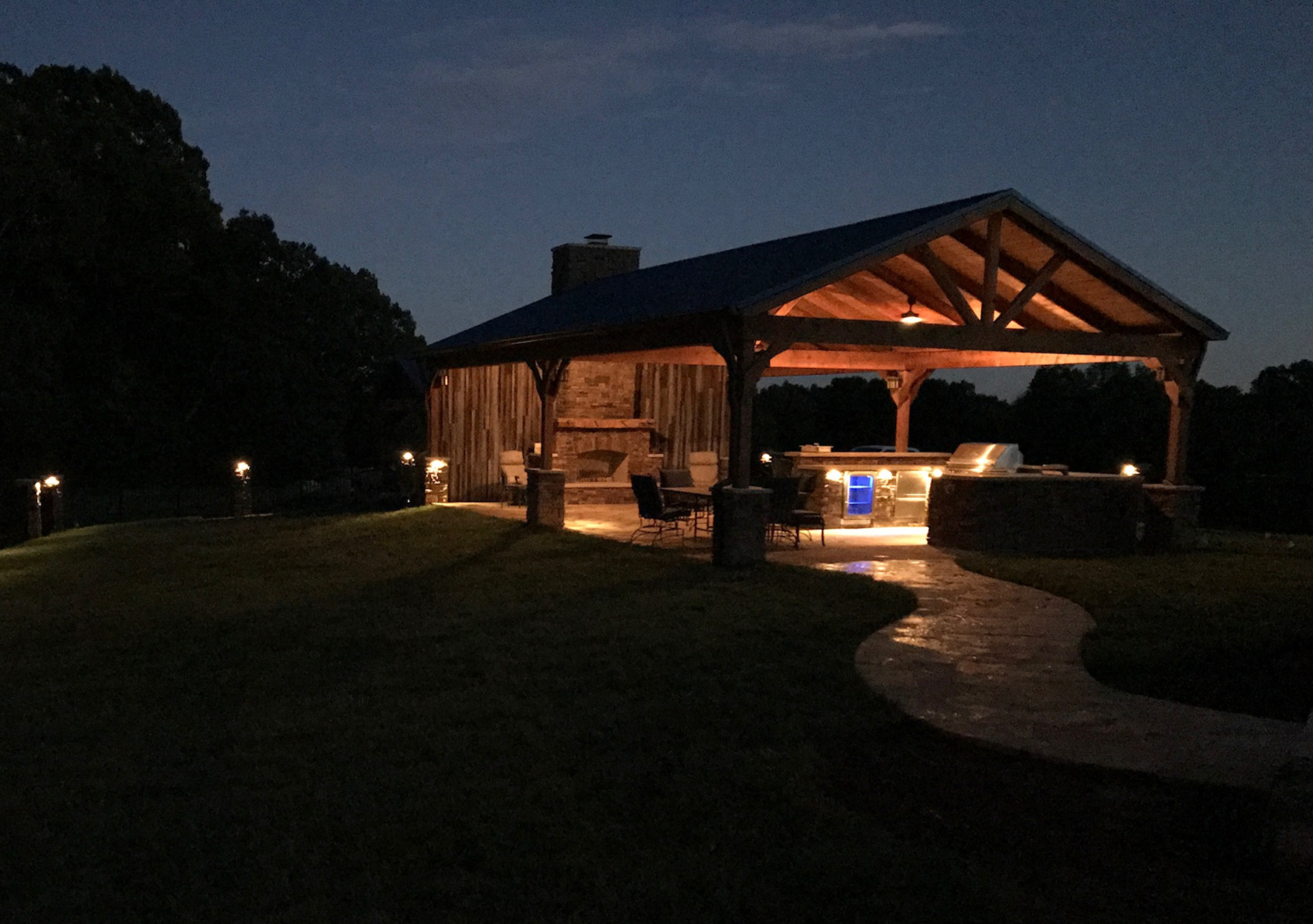 Freestanding timber pavilion with covered outdoor kitchen, stone fireplace, and landscape pathway lighting at dusk in Middle Tennessee